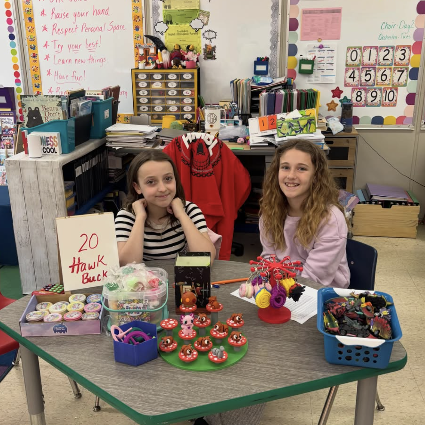 Two students sit behind a classroom table displaying prizes and rewards available through the Hawk Buck student store.